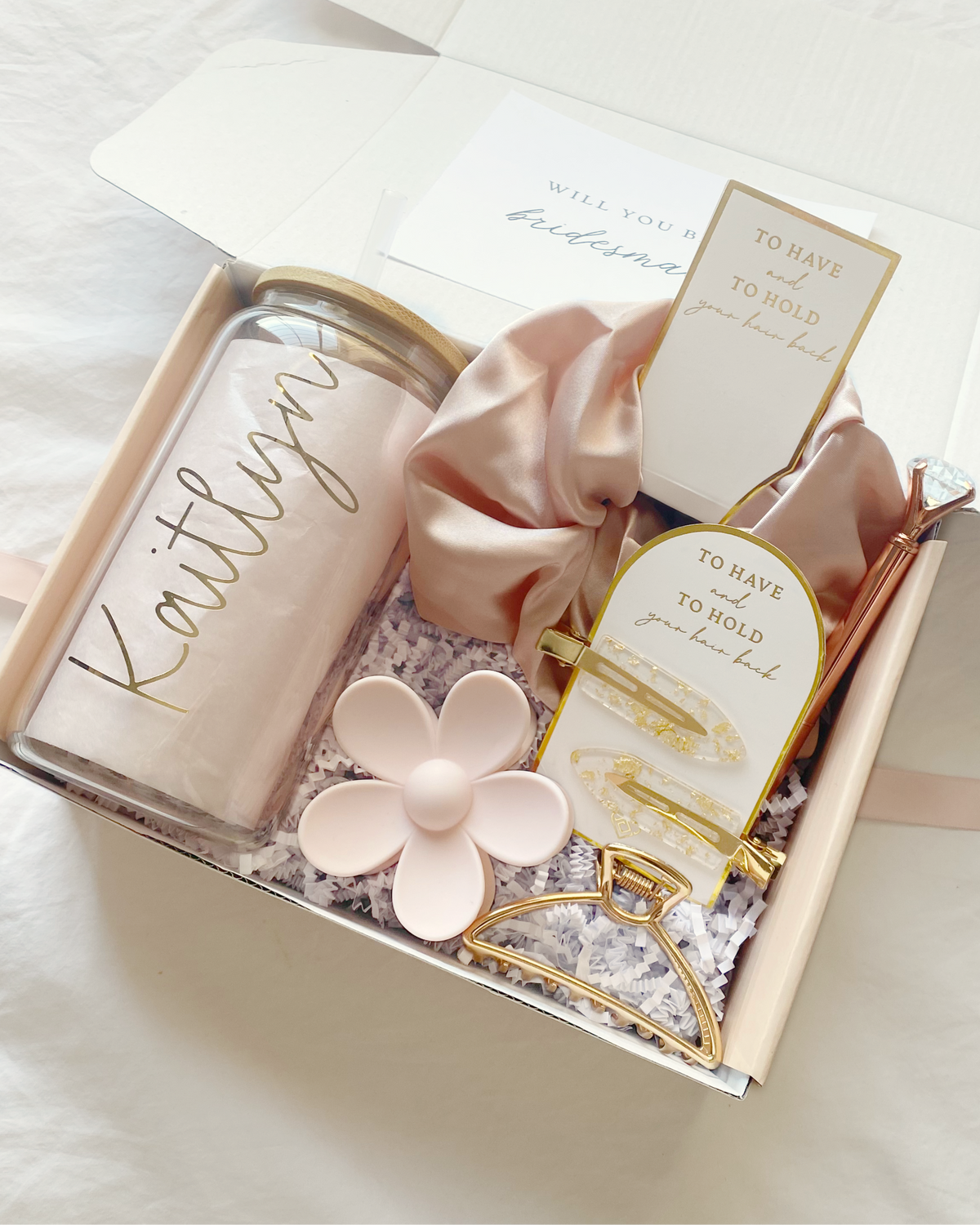 Personalized light pink bridesmaid gift set with customizable role titles – maid of honor, matron of honor, or your own message. Includes glass tumbler, jumbo scrunchie, chic gold claw clip, light pink daisy hair claw clip, pair of gold flake hair clips and diamond pen.