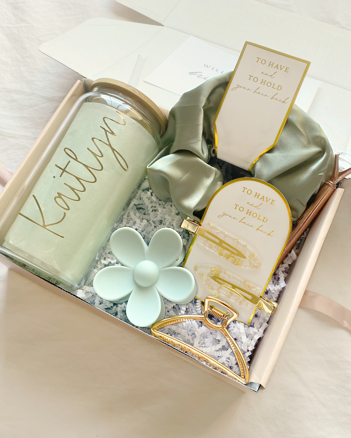 Personalized sage bridesmaid gift set with customizable role titles – maid of honor, matron of honor, or your own message. Includes glass tumbler, jumbo scrunchie, chic gold claw clip, sage green daisy hair claw clip, pair of gold flake hair clips and diamond pen.